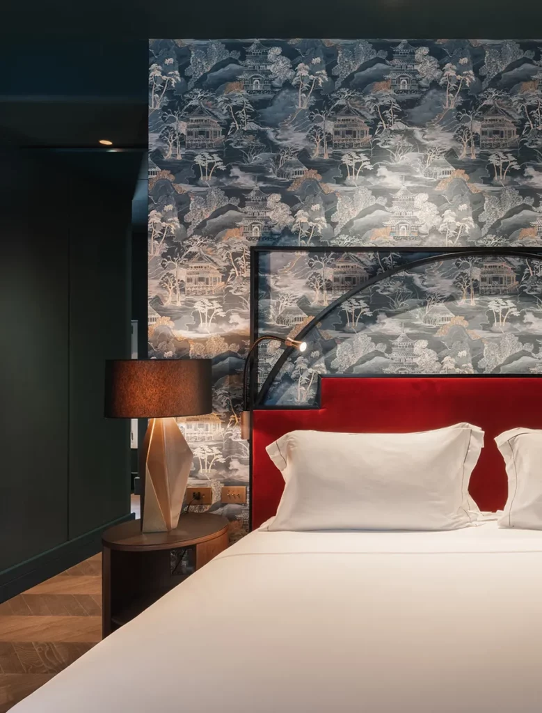 Testiera letto rossa con carta da parati orientale blu, lampada da tavolo scultorea e comodino in legno.